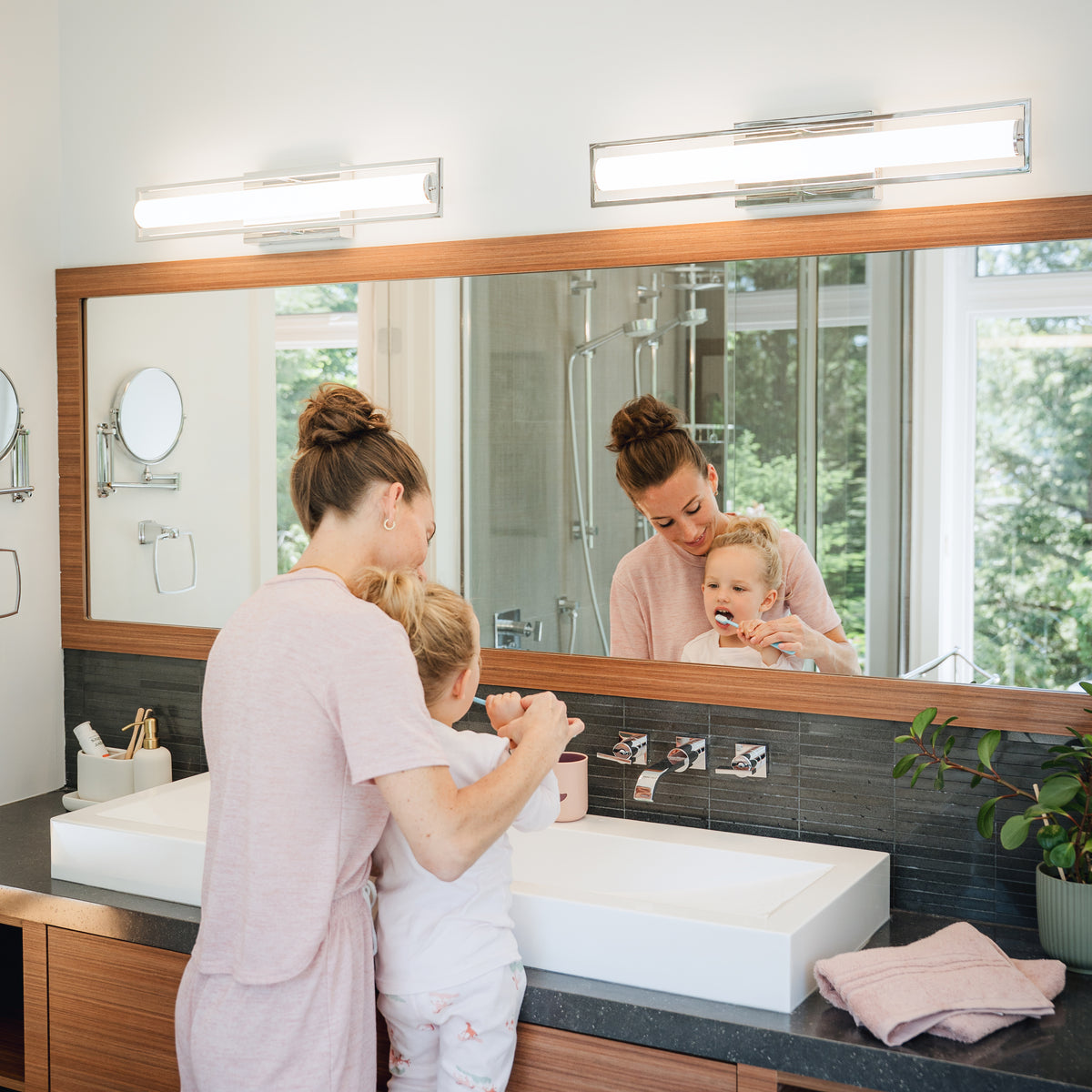 A woman and child brush teeth, smiling as the Artika Rialto 27-in LED Vanity Light Bar glows in the bathroom mirror.