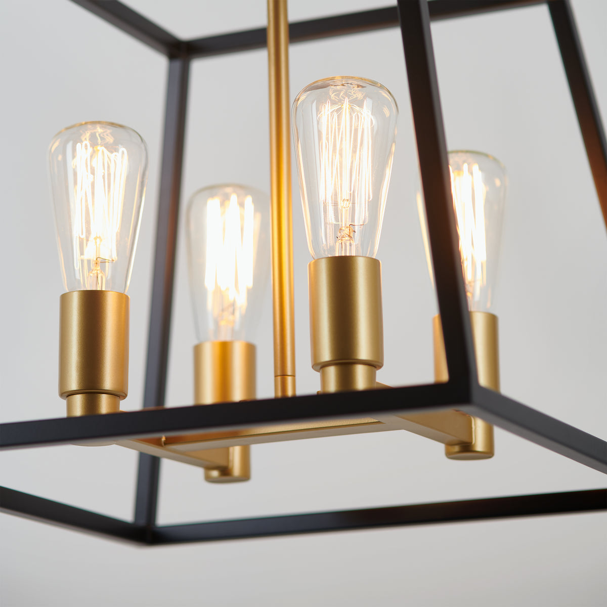 Close-up of the Artika Carter Square 4-light LED chandelier, featuring a geometric black metal frame, gold sockets, and exposed bulbs. The fixture is illuminated, casting a warm glow with modern industrial charm.