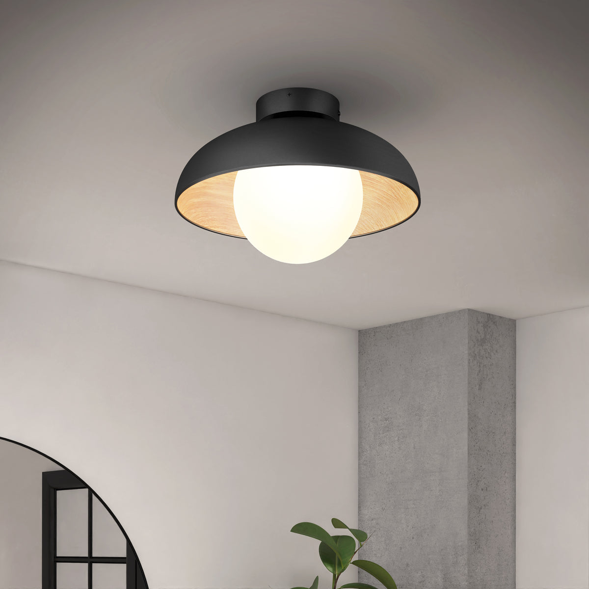 The Artika Boden LED flush mount light 5 CCT, a modern black ceiling fixture with a round white bulb, is mounted on a white ceiling above a gray concrete column, a leafy plant, and an arched window.