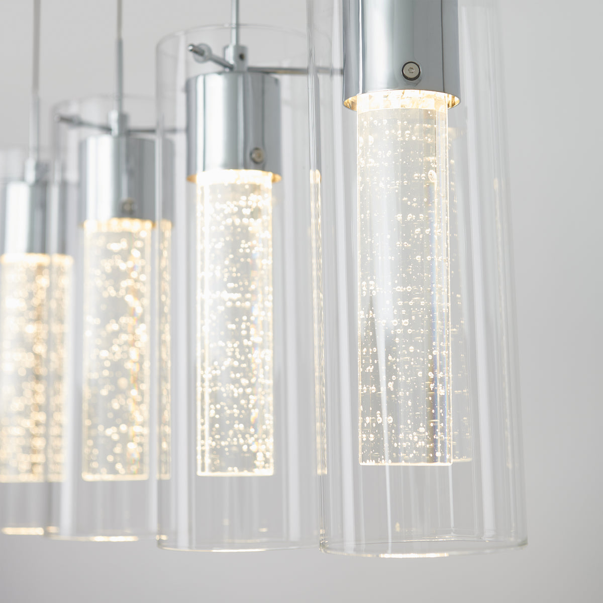 Close-up of the Artika Essence 4-light LED pendant, showcasing cylindrical clear glass with chrome accents and bubble-filled inner tubes, emitting a bright white glow against a plain background.