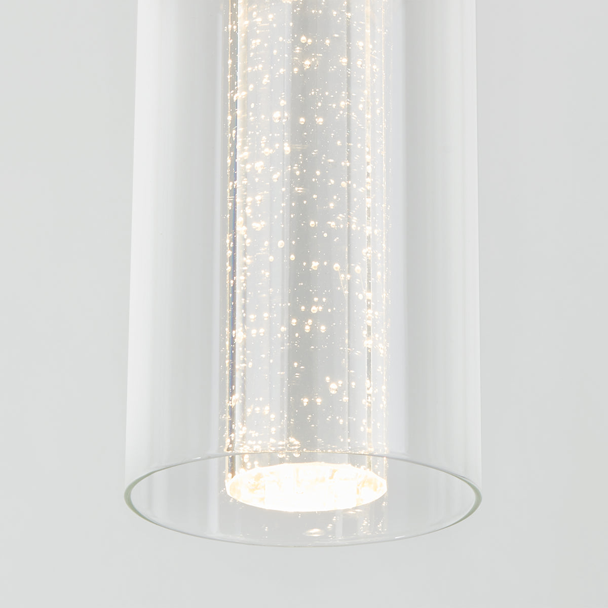 The Artika Bubble Flow 1-light pendant 2 features a modern cylindrical design with bubbles in the glass, casting a bright white glow against a plain light gray background.