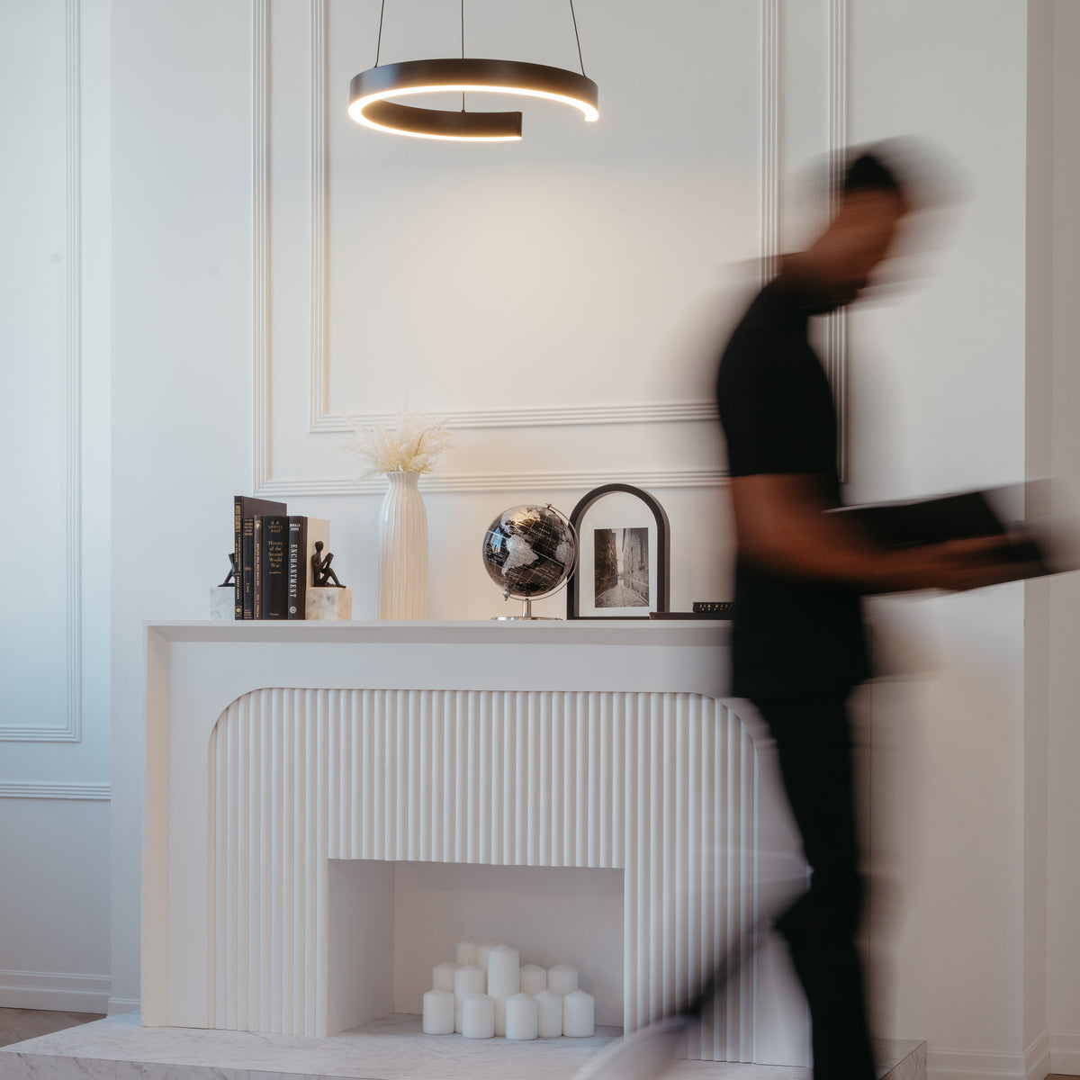 A blurred figure passes a modern white mantel adorned with books, a vase, globe, framed art, and candles. Overhead hangs the Artika Vance modern LED pendant light 3 CCT, illuminating the minimalist space.