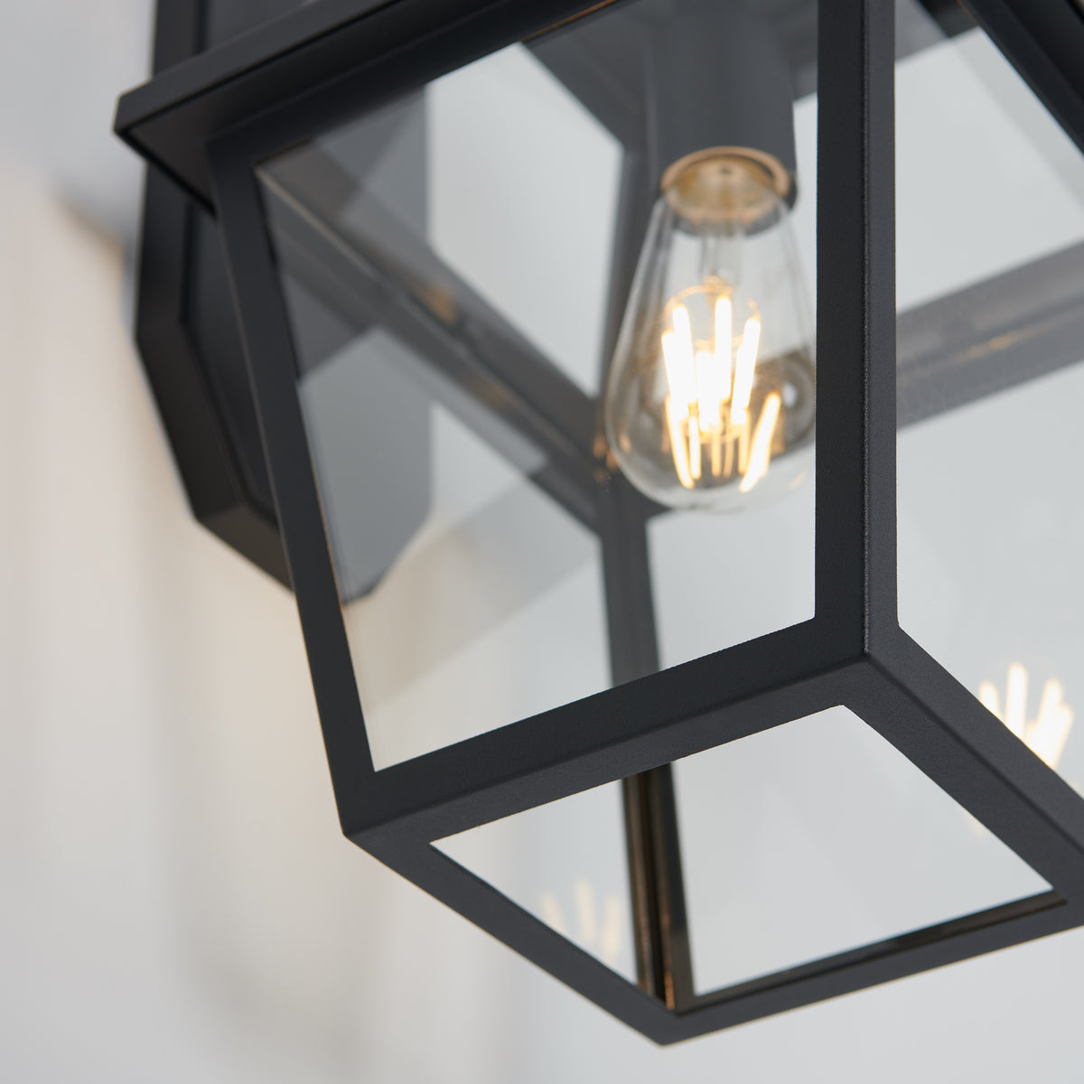 Close-up of the Artika Colonial outdoor lantern wall light, a modern black metal and glass fixture mounted on a wall with an exposed filament bulb glowing inside.