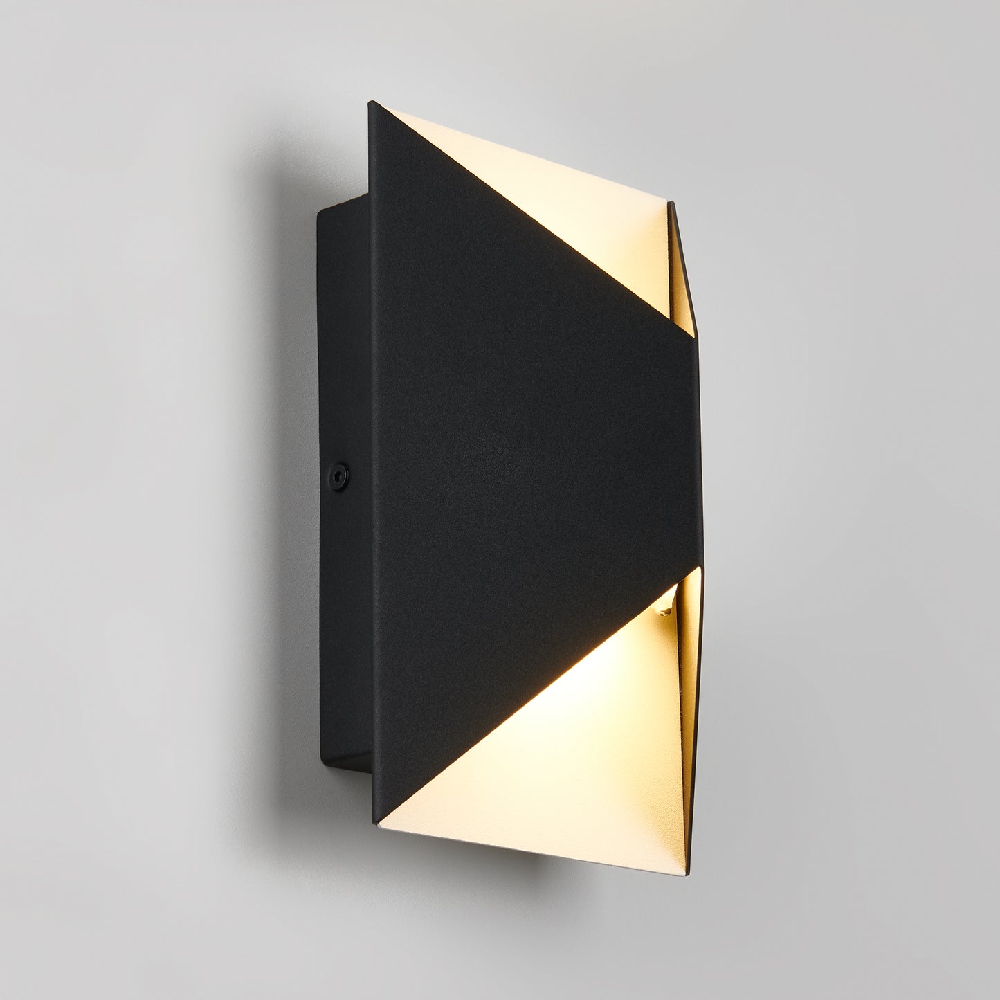 The Artika Tria LED indoor-outdoor wall light features a geometric black metal design with triangular openings at the top and bottom, casting soft, warm light onto a plain gray wall.
