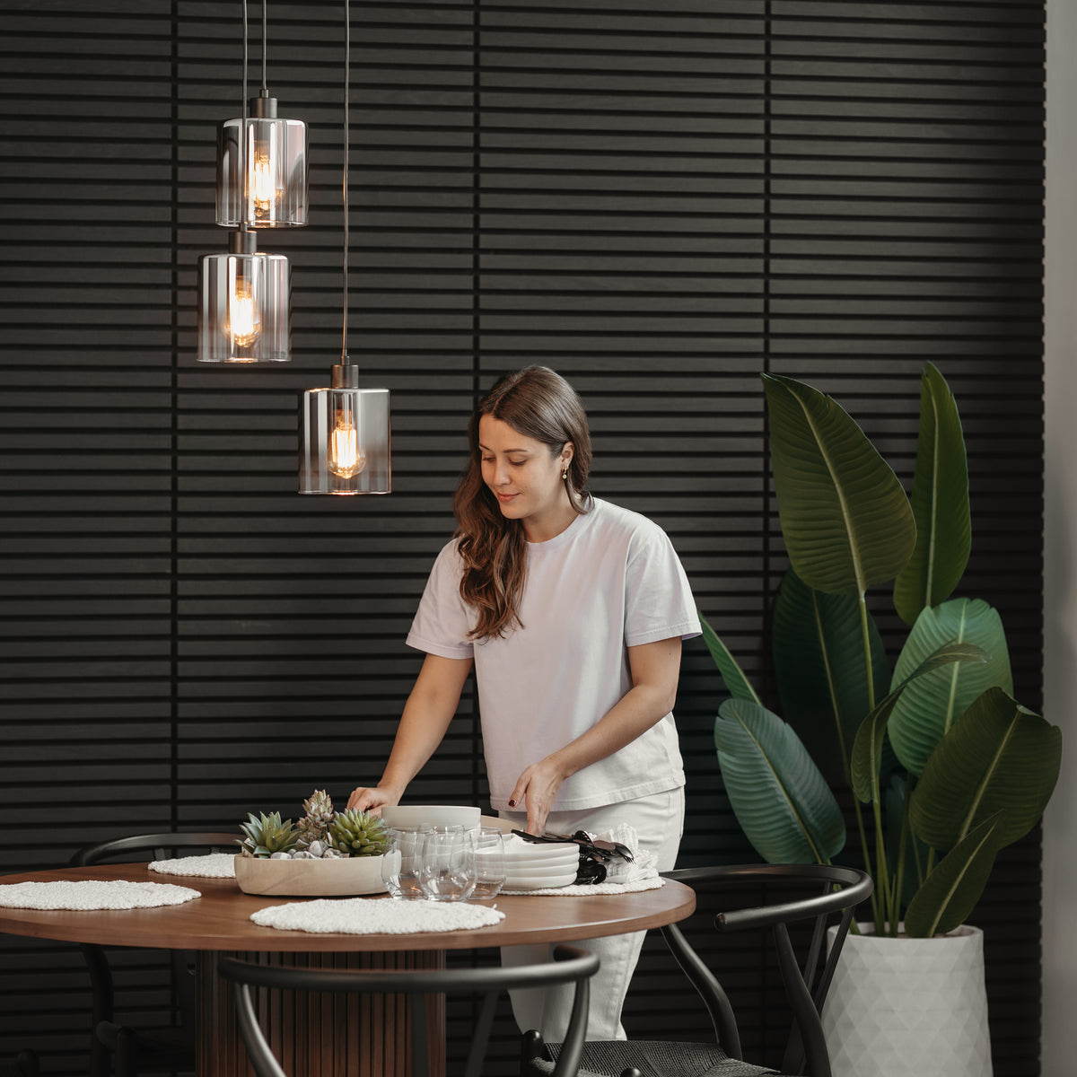 A woman in a white t-shirt sets a round dining table with placemats and glasses beneath the Artika Haze modern 3-light pendant light, with a large potted plant and a black paneled wall in the background.