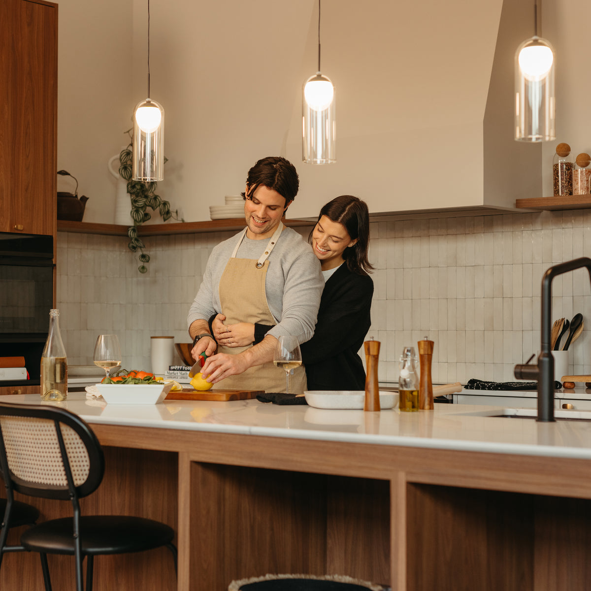 A couple enjoys cooking in a cozy, well-lit kitchen illuminated by the Artika Mirah LED modern mid-century pendant light 3 CCT as one cuts a yellow bell pepper and the other hugs from behind.