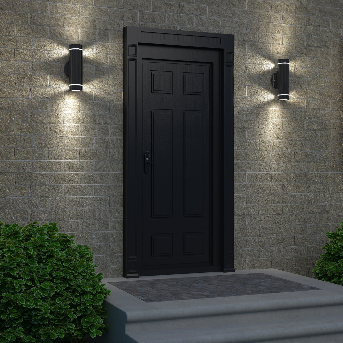 A black front door with decorative panels sits in a beige brick wall, flanked by two Artika C7 indoor-outdoor wall lights. Two small green bushes frame the entry, and a stone step leads up to the door.