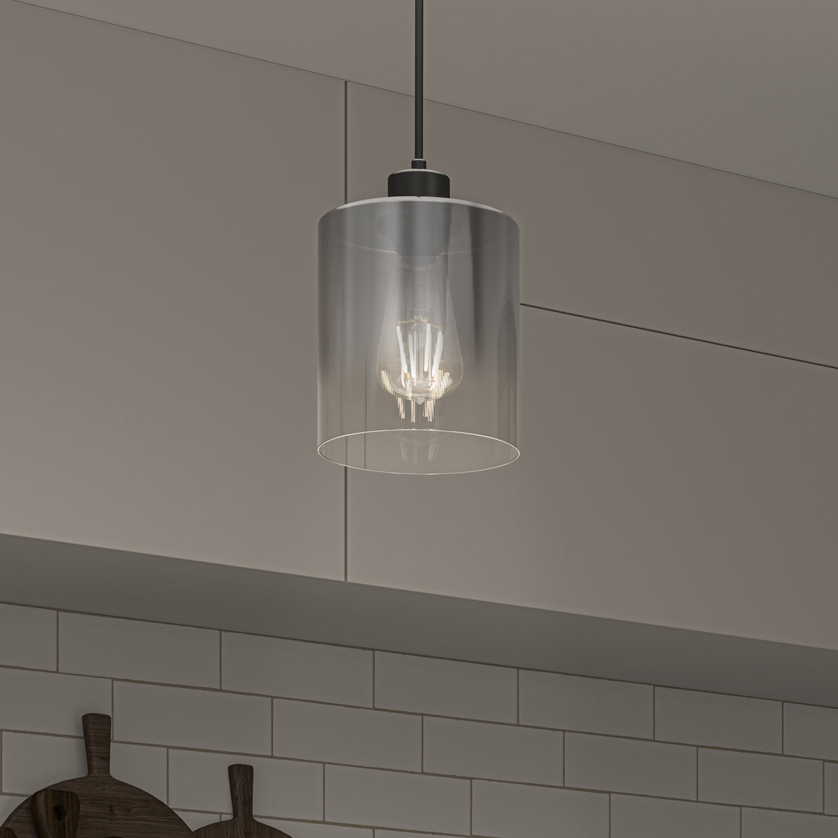 The Artika Haze pendant light with a clear glass shade and visible bulb hangs from the ceiling in a kitchen with gray cabinets and a white subway tile backsplash.