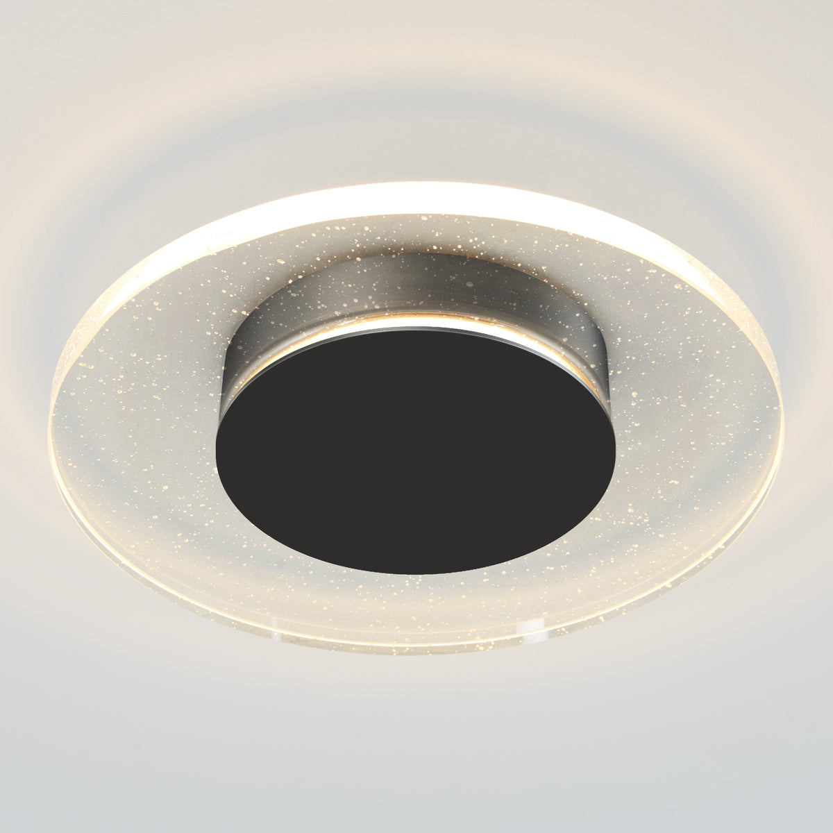 The Artika Essence Disk LED flush mount is a modern ceiling light featuring a black center, transparent outer ring with sparkling specks, and emits a soft white glow.