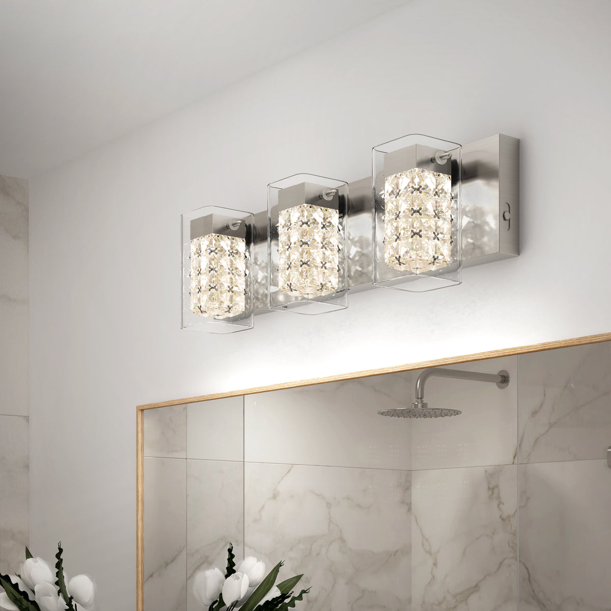 The Artika Crystal Cube LED bathroom vanity light 5 CCT features three crystal-like square lights above a large mirror, reflecting marble-patterned walls and a rainfall showerhead. White flowers sit in the bottom left corner.
