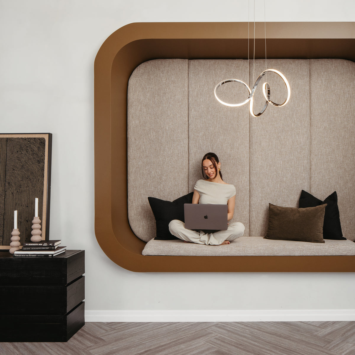 A woman sits cross-legged on a modern built-in alcove seat with neutral cushions, using a laptop. Overhead, the Artika Swirl LED pendant light adds style, while a side table holds candles and framed art for a cozy, contemporary vibe.