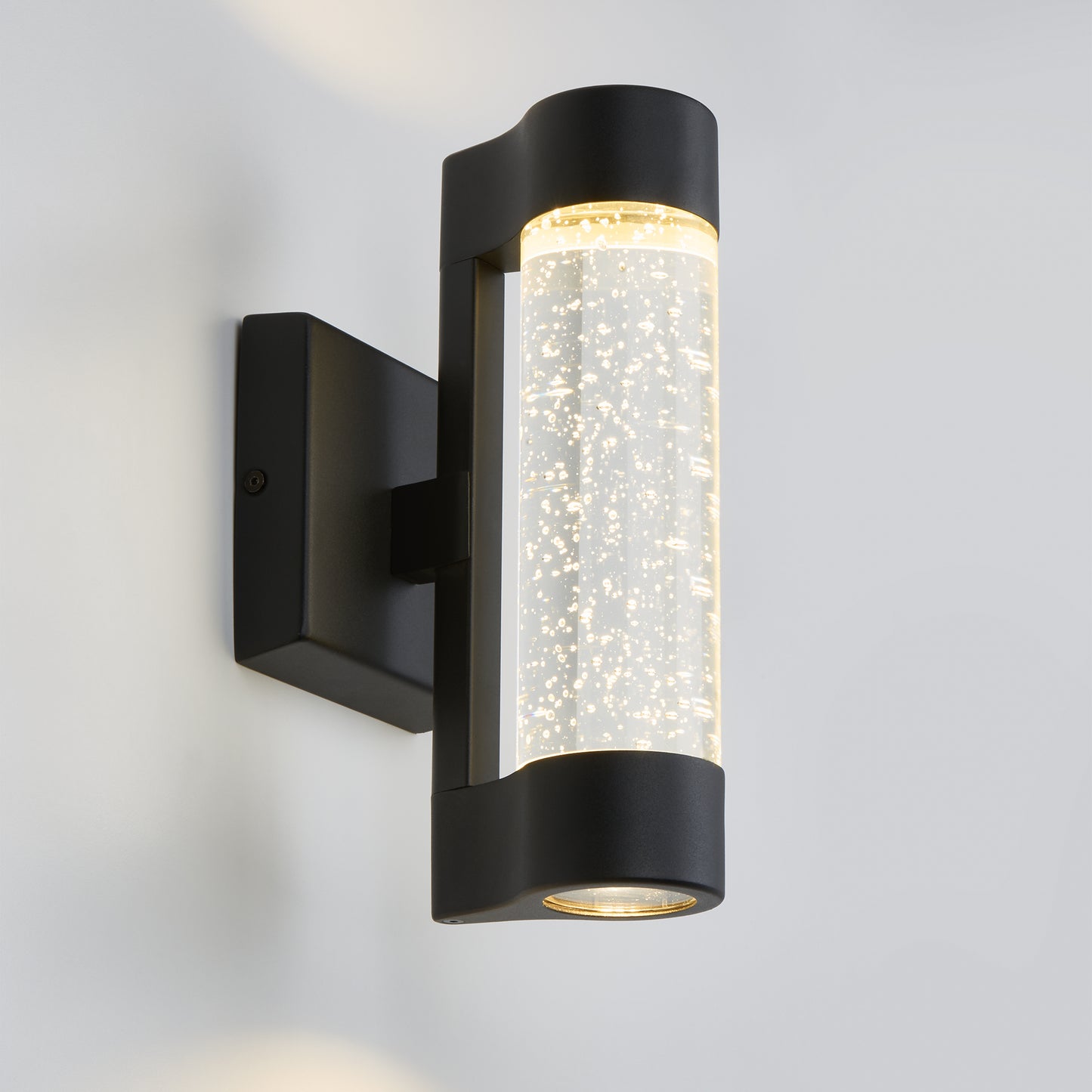 The Artika Bubble Element LED indoor-outdoor light features a modern black design with a bubble-filled cylindrical glass shade, casting a warm glow against any white wall.