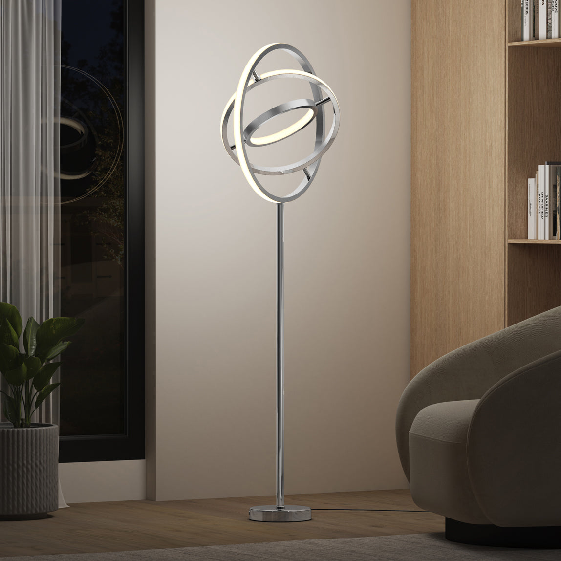 The Artika Pivot modern LED floor lamp 3 CCT, featuring circular ring-shaped lights, stands in a contemporary living room beside a curved beige chair, bookshelf, potted plant, and large window.