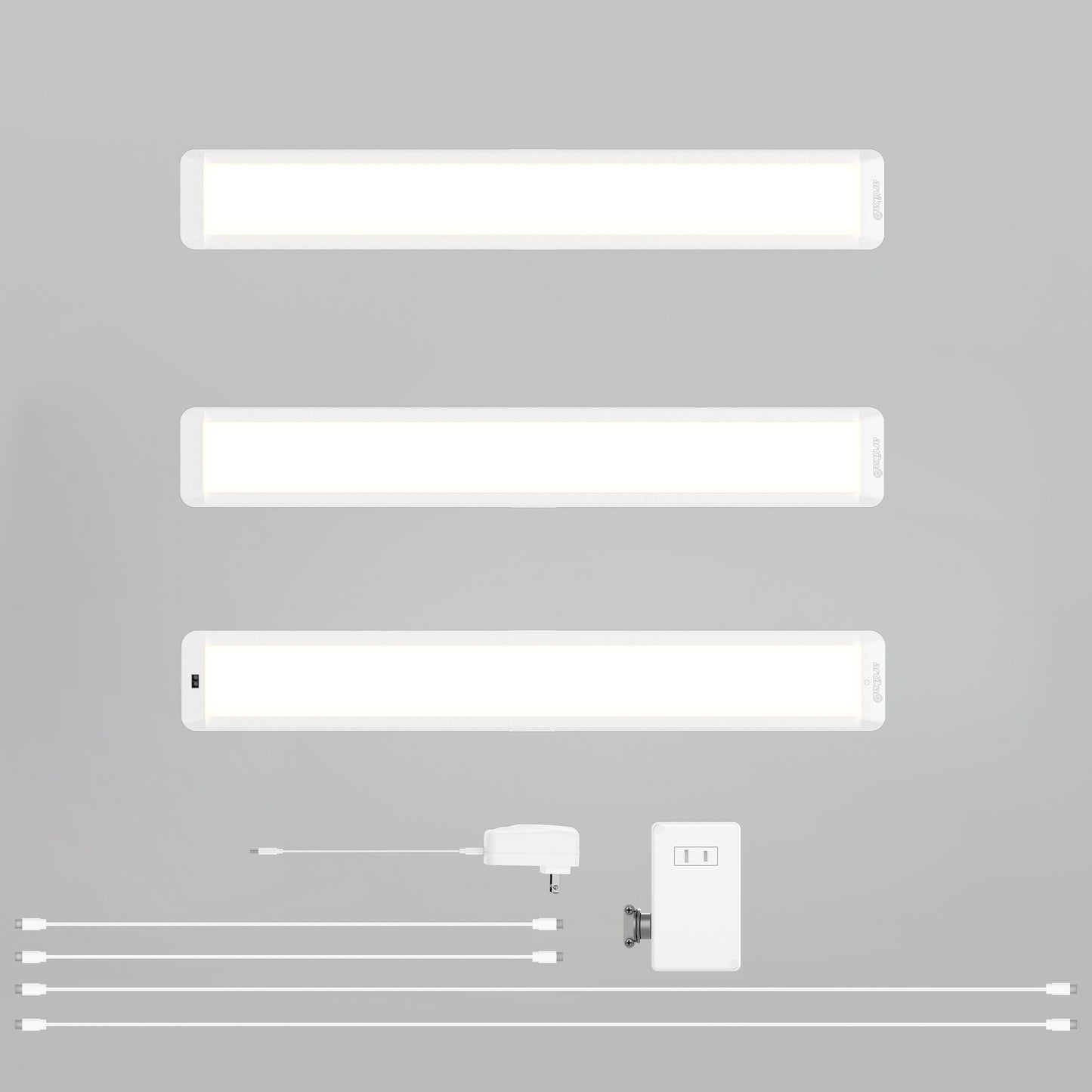 Three Artika Maestro under cabinet LED light 3CCT bars are shown horizontally above accessories: four cables, a power adapter, a switch box, and a mounting bracket, all set on a plain gray background.