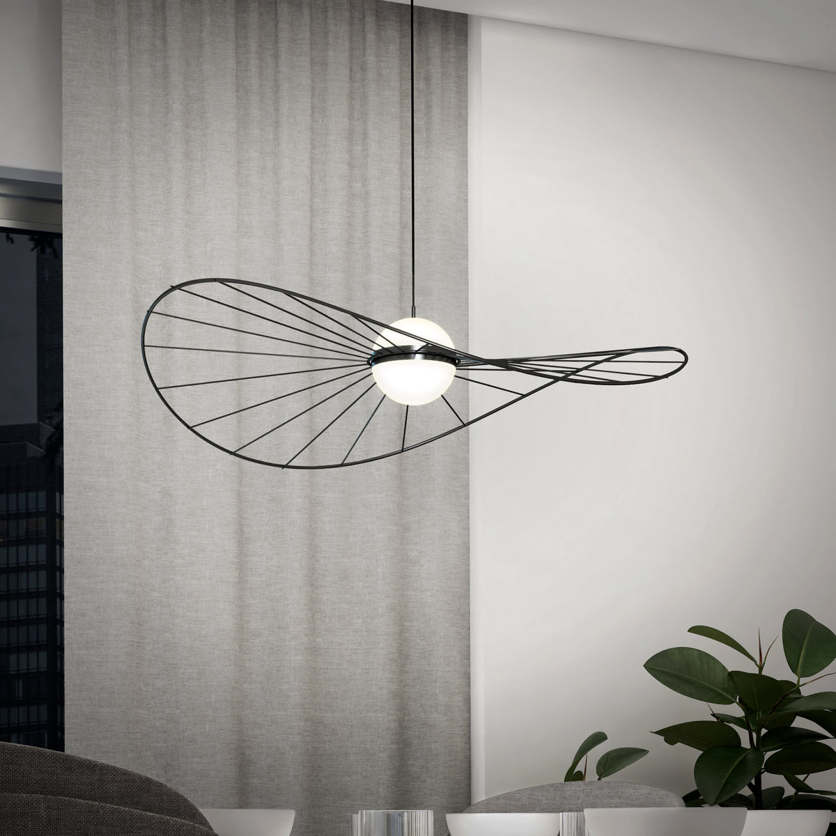 The Artika Tolena LED pendant light 3 CCT, featuring a sculptural black wire frame and two white globes, hangs in a minimalist room with light gray curtains, a window, and a green indoor plant.