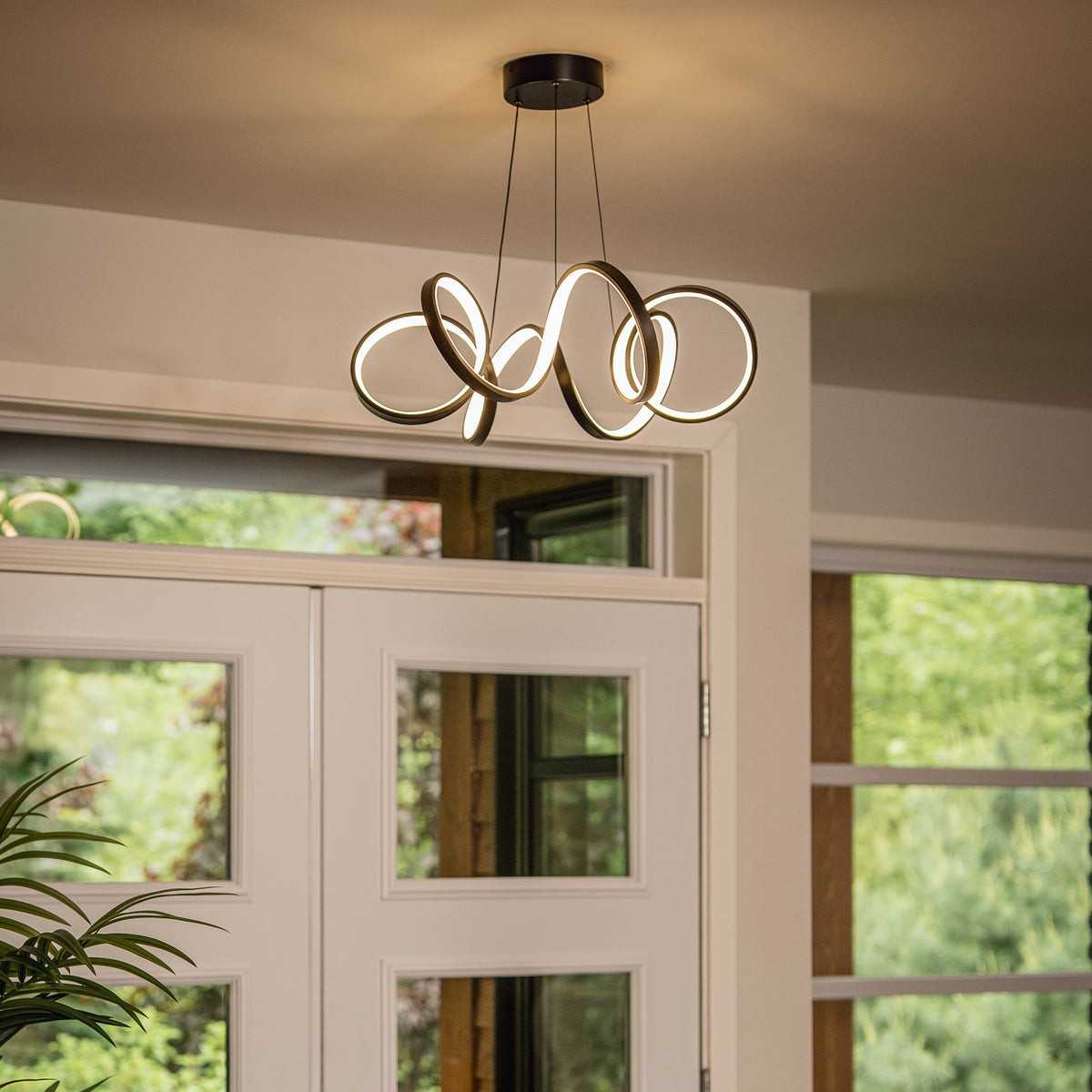 Artika Swirl Ribbon LED pendant light hangs by white doors, with greenery seen through nearby windows.