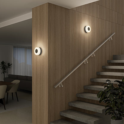 A modern interior features a staircase with stone steps, wood-paneled walls, and two Artika Ludwig modern LED indoor-outdoor wall lights. A handrail runs beside the stairs, and a dining area with table and chairs is visible in the background.
