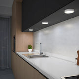 Modern kitchen with a sleek white countertop, stainless steel sink, faucet, under-cabinet lighting, a small potted plant, wooden paneling, and a pepper grinder near a stovetop.