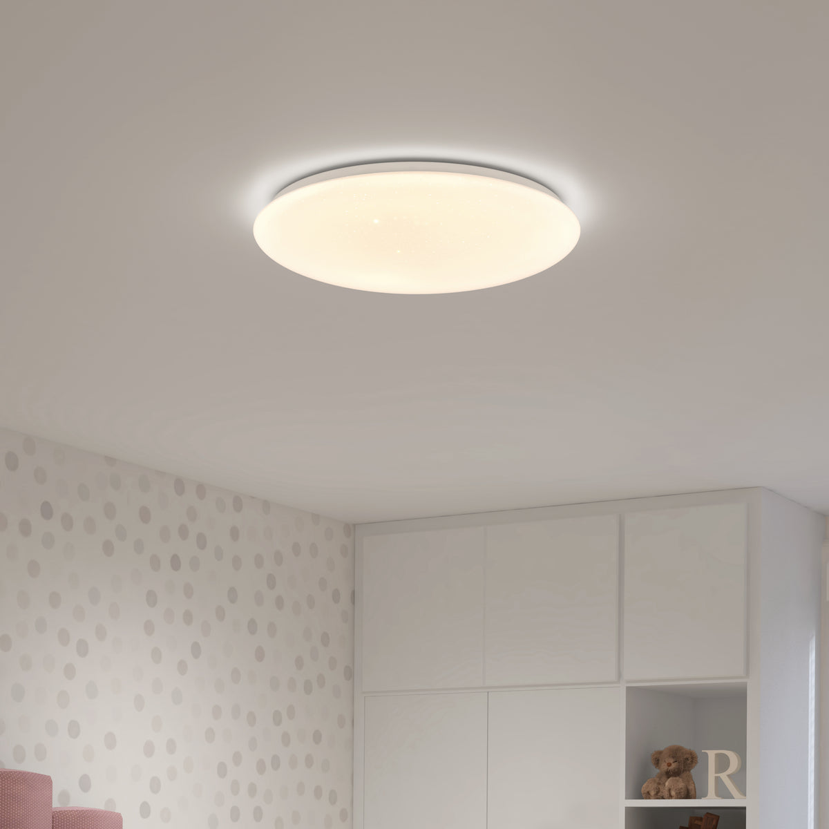 A round, modern ceiling light glows softly in a room with white walls featuring a subtle polka dot pattern. White cabinets and shelves, some holding a teddy bear and a letter R, are visible below.