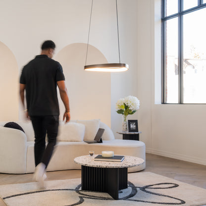 A man walks through a modern living room, illuminated by the Artika Bjorn modern LED pendant light (5 CCT), featuring a white curved sofa, round coffee table, side table with flowers, and large windows with natural light.