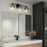 A modern bathroom with a large mirror, wooden vanity, black faucet, and a three-light fixture with glass shades above the mirror. A vase with dried plants sits on the white countertop. Dark tiles cover one wall.