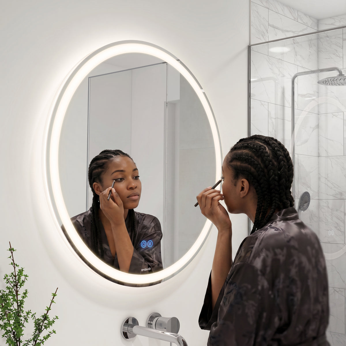 A woman with braided hair applies eyeliner while using the Artika Ayla 28 in. LED mirror 3 CCT with defogger in a modern bathroom with a glass shower, small potted plant, and adjustable lighting.