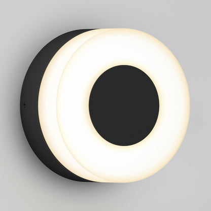 The Artika Ludwig modern LED indoor-outdoor wall light 5CCT features a black circular center with a glowing white ring, creating a sleek, contemporary look when mounted on a light gray wall.