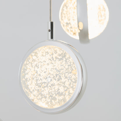 Close-up of the Artika Avalon 4-light LED pendant, featuring a modern circular design, transparent center with bubbles, and a bright warm white glow. Another Avalon pendant appears blurred in the background.