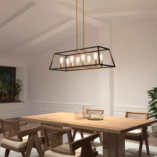 A modern dining room with a wooden table, four woven chairs, white walls, plants, and a window lit by evening light features the Carter Long 8-light Chandelier by Artika—a rectangular black and gold fixture with exposed bulbs.
