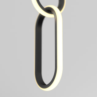 A close-up of Artikas Catena LED pendant light 5 CCT shows two interlocking, oval rings with a modern black finish, emitting a soft white edge glow, set against a plain light gray background.