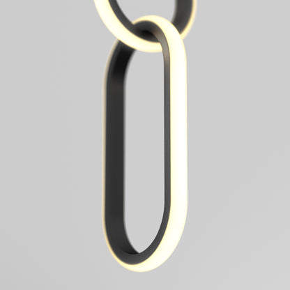 A close-up of Artikas Catena LED pendant light 5 CCT shows two interlocking, oval rings with a modern black finish, emitting a soft white edge glow, set against a plain light gray background.