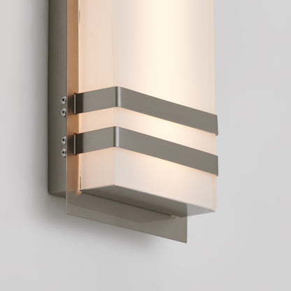 A modern wall-mounted light fixture with a frosted rectangular shade and two horizontal metal bands, emitting a soft, warm glow against a plain white wall.