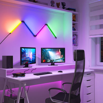 A modern gaming setup featuring dual monitors on a white desk, an ergonomic black chair, shelves with figurines and books, and the Artika Fitch smart foldable RGB LED gaming floor lamp for colorful ambient lighting.