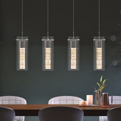 The Artika Bubble Flow 4-light pendant, featuring cylindrical glass shades, hangs above a wooden dining table adorned with candles, plants, and a copper carafe. Light gray upholstered chairs and a dark green wall complete the scene.