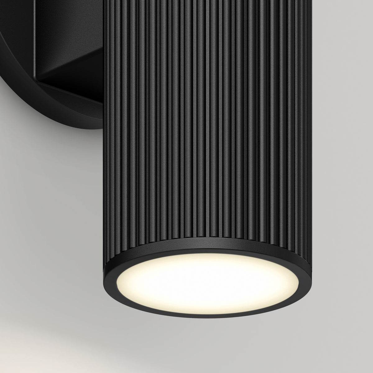 Close-up of the Artika Groove LED indoor-outdoor wall light, featuring a modern black cylindrical design with vertical ridges. Mounted on a wall, it emits a soft white downward glow.