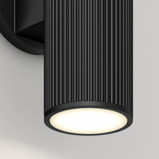 Close-up of the Artika Groove LED indoor-outdoor wall light, featuring a modern black cylindrical design with vertical ridges. Mounted on a wall, it emits a soft white downward glow.