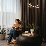A woman sits in a modern armchair by a window, reading a book. She wears jeans, a sleeveless top, and slippers. Nearby are plants, books, a candle, and a round black table. Soft natural light fills the room.