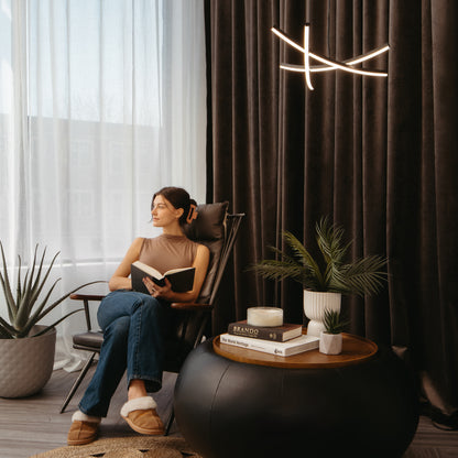 A woman relaxes in a modern chair by the window, reading beside potted plants and books, with a lit candle atop a round black table. Behind her hang dark curtains and an Artika Orly modern LED pendant light (5 CCT).