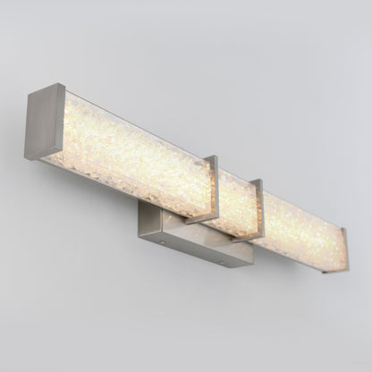 The Artika Royale LED bathroom vanity light features a brushed metal base and three rectangular, crystal-embedded panels, casting a soft warm glow when mounted on a light gray wall.