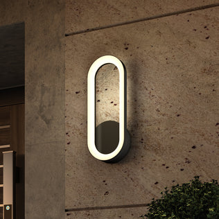 The Artika Aiden modern LED indoor-outdoor wall light 5 CCT, with an oval shape and glowing border, is mounted on a beige stone wall beside a door, with a green shrub visible in the lower right corner.