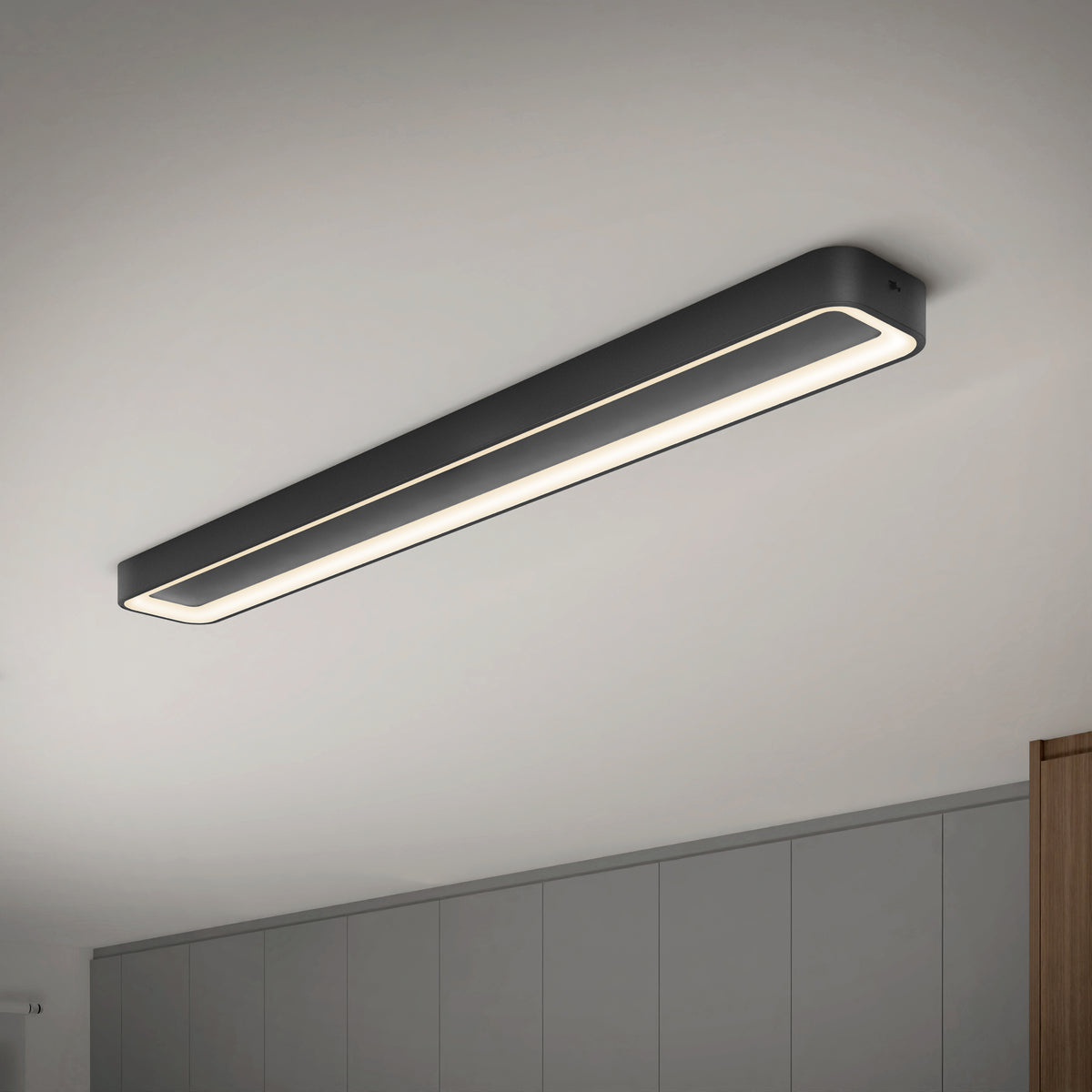A modern, rectangular black ceiling light fixture with a continuous LED strip is mounted on a white ceiling above gray cabinets in a minimalist room.