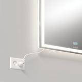 A close-up of a wall-mounted mirror with LED lights around its edge. A white power cord is plugged into a nearby wall socket below the mirror. The mirror displays faint touch controls on its surface.
