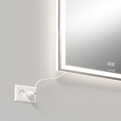 The Artika Aurea anti fog LED wall mirror hangs above a power outlet, powered by a white cord. Both the sleek mirror with its glowing frame and the wall feature a clean, minimalist design.