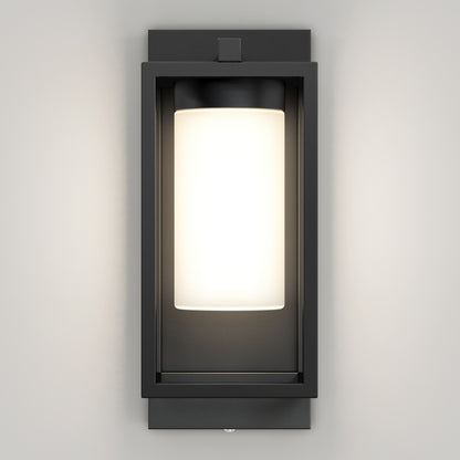 The Artika Aurel Outdoor LED Light, 3CCT features a rectangular black metal frame with a frosted cylindrical glass shade, mounted on a light wall and illuminated to cast a gentle glow.