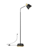 A modern black floor lamp with a gold accent, angled stem, and conical shade stands on a round base. The lamp is plugged into a wall outlet with a visible black cord and foot switch.