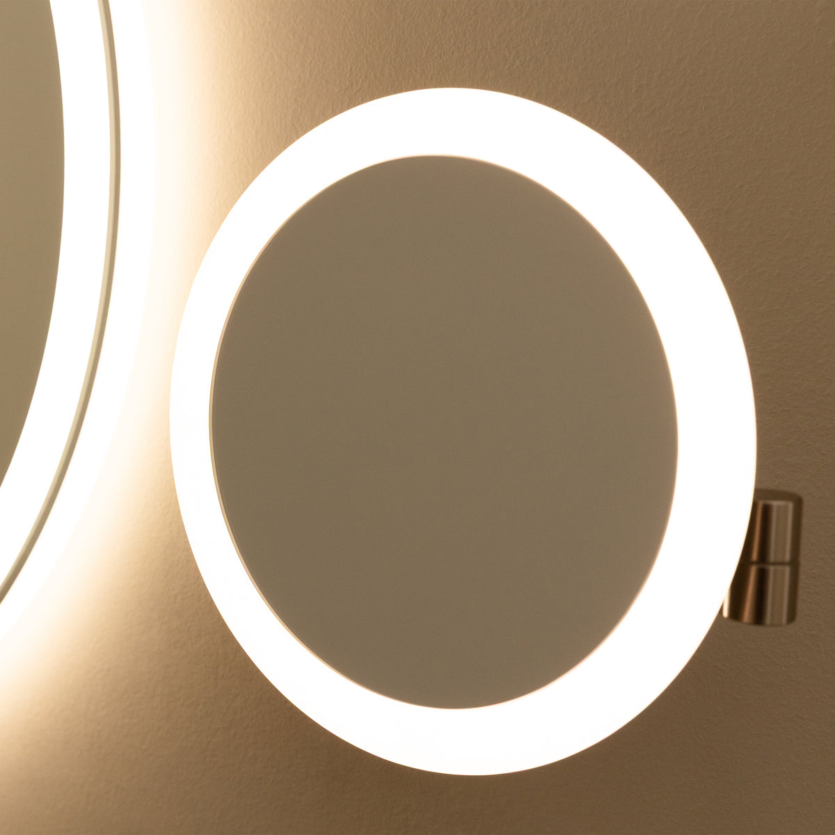 A round, wall-mounted mirror with a bright, illuminated LED light ring around its edge, reflecting a portion of another lit mirror on a neutral-colored wall.