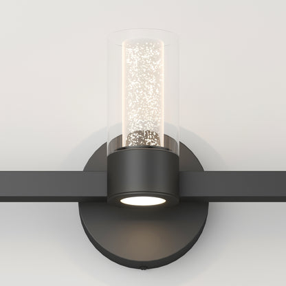 The Artika Novello modern LED bathroom vanity light features a black circular base, horizontal arm, and a cylindrical bubble glass shade around a central light, shown mounted on a white wall.