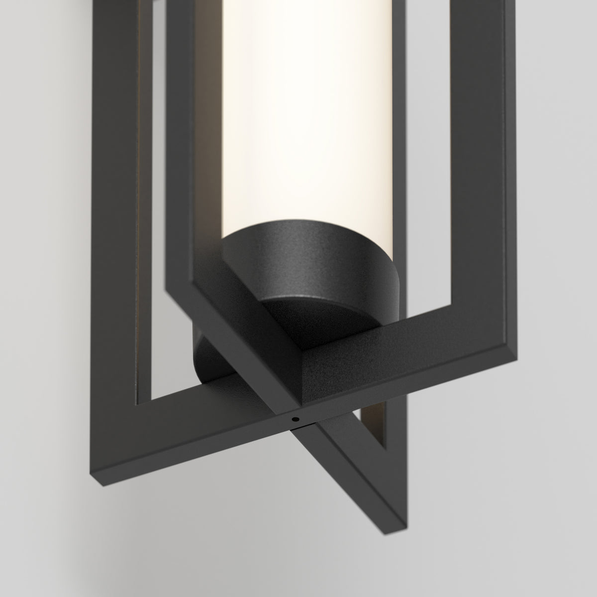 Close-up of a modern wall sconce featuring a black geometric metal frame surrounding a cylindrical frosted glass light, mounted on a light gray wall.