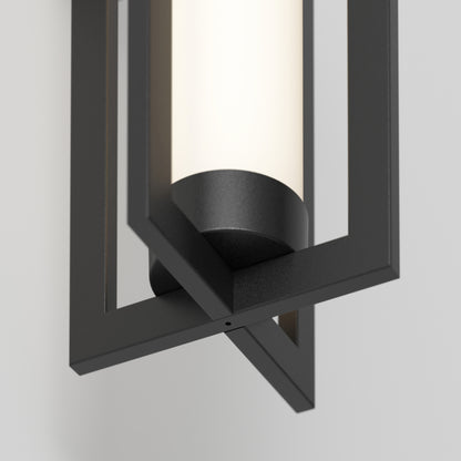 A close-up of the Artika Walt indoor-outdoor LED wall light shows its cylindrical frosted glass within a black geometric frame, mounted on a light-colored wall.