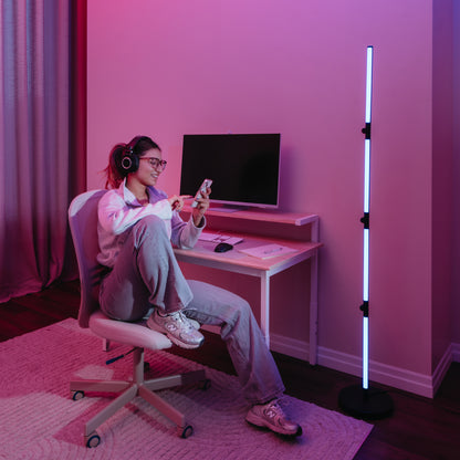 A woman wearing headphones and glasses sits comfortably in a modern room with pink-purple lighting, using her smartphone. Beside her is the Artika Fitch smart foldable RGB LED gaming floor lamp near a desk with a computer setup.